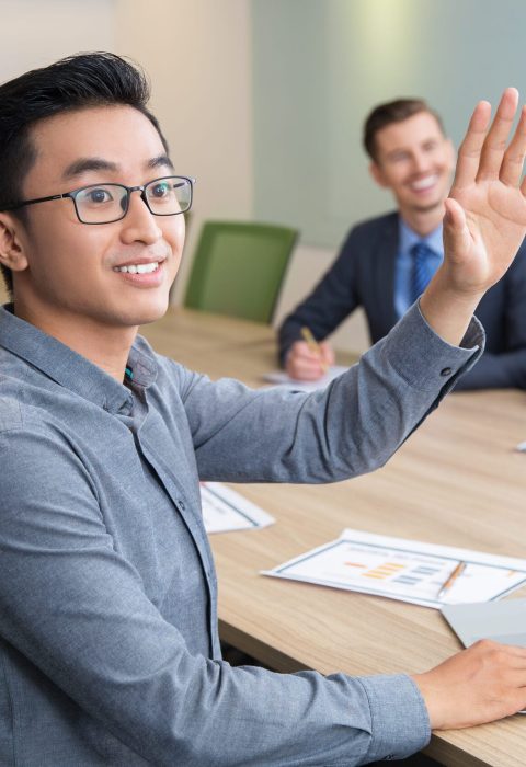 Young Asian businessman wearing glasses sitting at meeting or conference in boardroom with multiethnic colleagues and raising hand to ask question